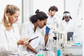 Several students in white coats work with microscopes and other equipment in a lab