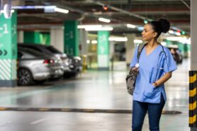 Healthcare worker dressed in scrubs and jeans looks for a car in a hospital garage