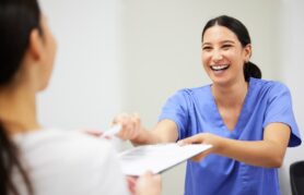 Smiling dental assistant hands patient forms to fill out before appointment