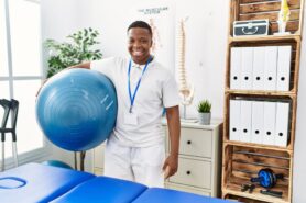 Smiling physical therapist assistant holds exercise ball in physical therapy office