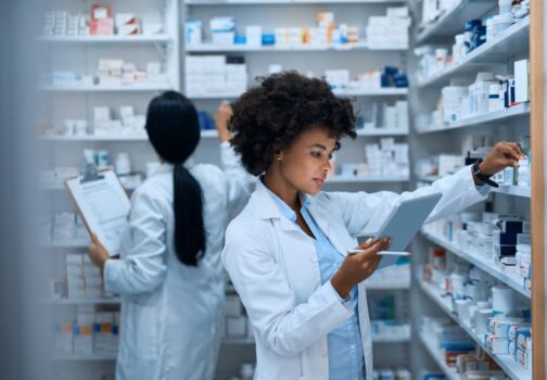 Pharmacy technician doing inventory in pharmacy with digital tablet and colleague in background